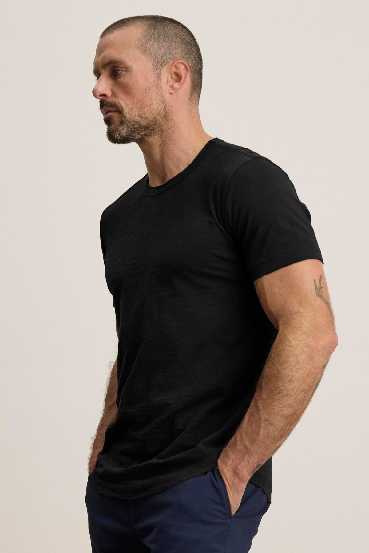 A bearded man with short hair wears the AMARO TEE by Velvet by Graham & Spencer, a black t-shirt crafted from lightweight Peruvian cotton with a classic crew neck, paired with blue pants. He's standing against a plain background with his hands in his pockets.-38516739342529