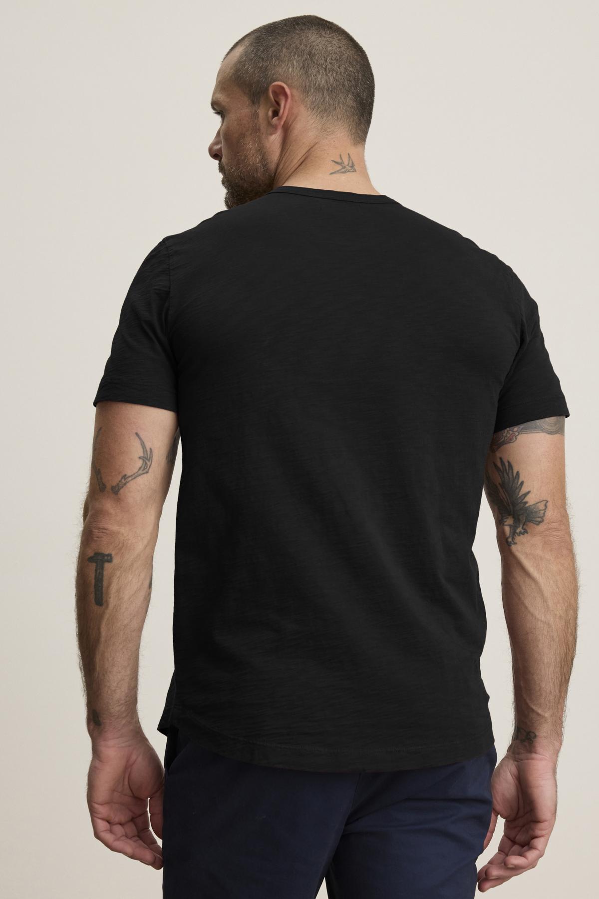   A man with tattoos, facing away, wears the AMARO TEE by Velvet by Graham & Spencer. This black t-shirt features a classic crew neckline and lightweight Peruvian cotton, pairing perfectly with dark pants against a neutral backdrop, making it an ideal layering piece. 