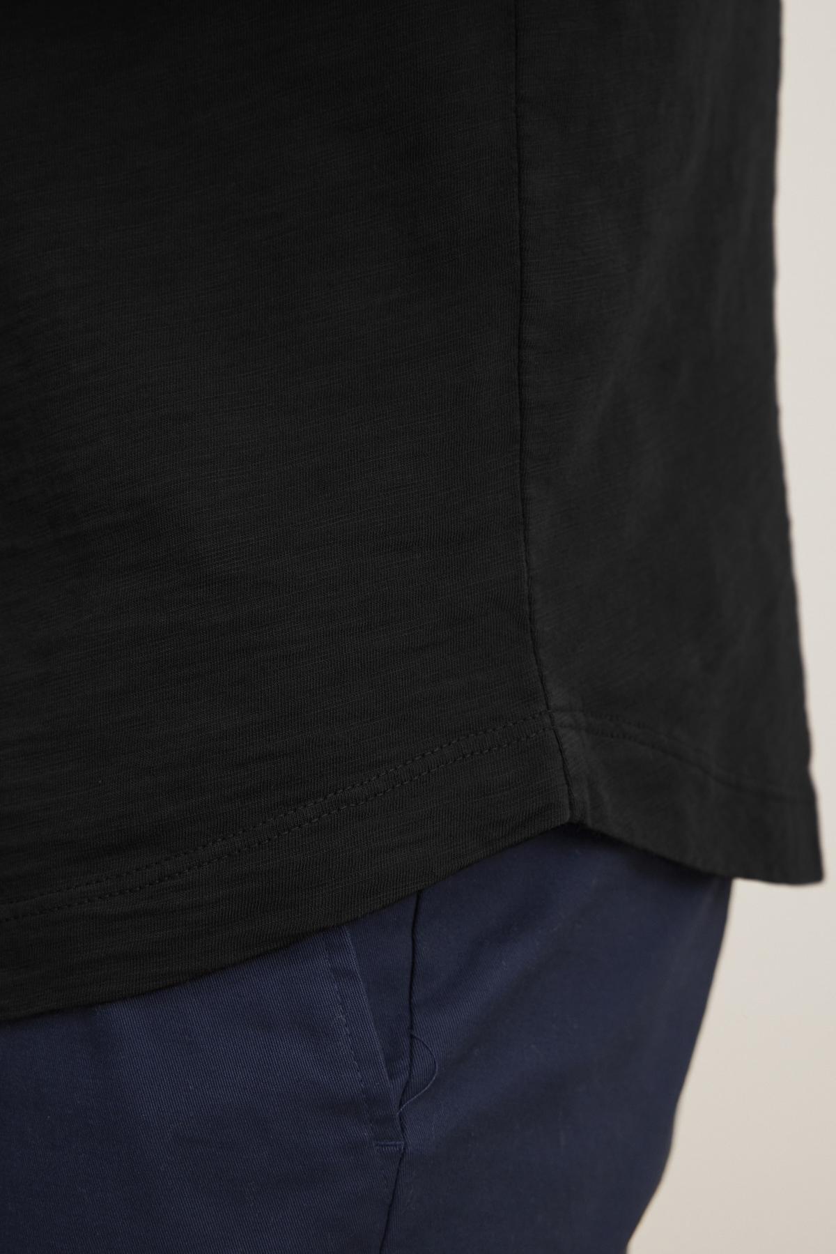   Close-up of someone wearing the AMARO TEE by Velvet by Graham & Spencer, a black shirt with a classic crew neckline crafted from lightweight Peruvian cotton, paired with blue pants showcasing the upper section. 