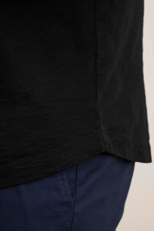 Close-up of someone wearing the AMARO TEE by Velvet by Graham & Spencer, a black shirt with a classic crew neckline crafted from lightweight Peruvian cotton, paired with blue pants showcasing the upper section.