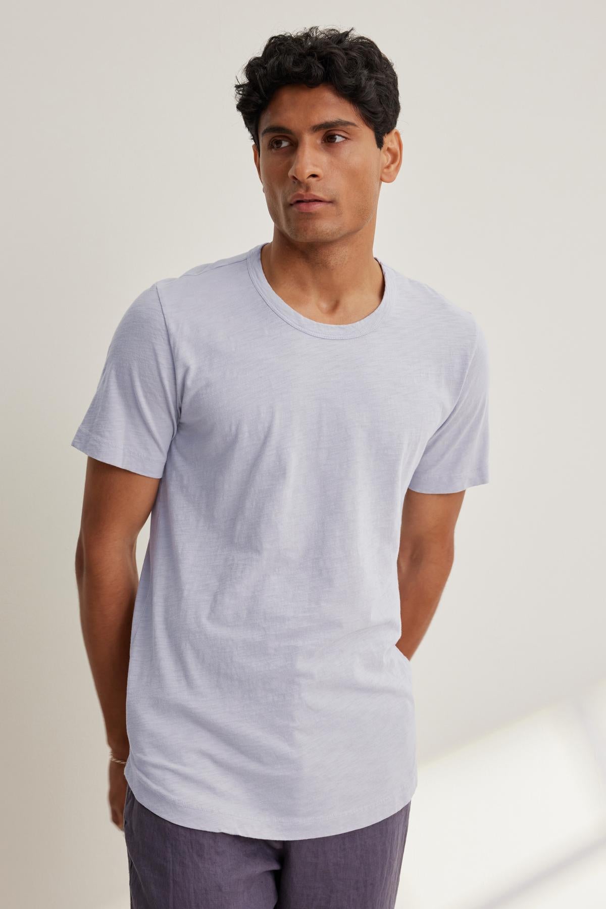 A person in the AMARO CREW NECK SLUB TEE by Velvet by Graham & Spencer, light gray and paired with dark pants, stands with hands behind their back, presenting a versatile look while gazing to the side against a plain background.-38658653487297