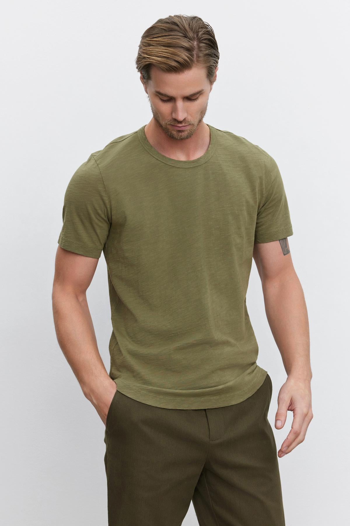   A man with short hair wearing the AMARO TEE, an olive green Peruvian cotton t-shirt by Velvet by Graham & Spencer, with a classic crew neckline and brown pants stands against a plain background, looking down. 