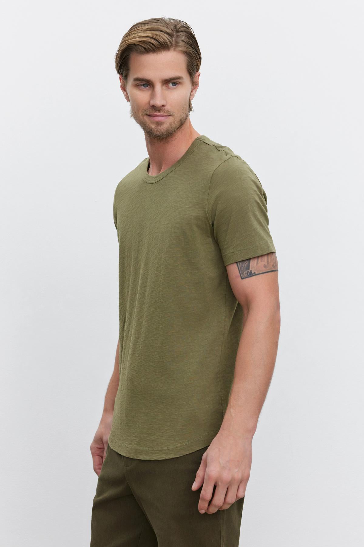   A man with light hair wearing an olive green AMARO TEE by Velvet by Graham & Spencer, made from lightweight Peruvian cotton, along with matching pants stands against a plain white background. 