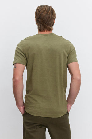 A person with light brown hair wearing an olive green AMARO TEE by Velvet by Graham & Spencer and dark green pants is standing with their back to the camera.