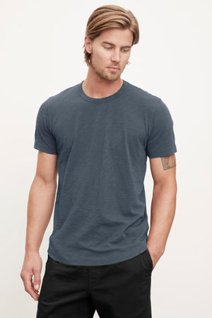 A person with long hair wearing a relaxed stylish feel, casual, short-sleeved dark gray Velvet by Graham & Spencer AMARO TEE and black pants stands against a plain light background. They have a neutral expression and a tattoo on their left arm.