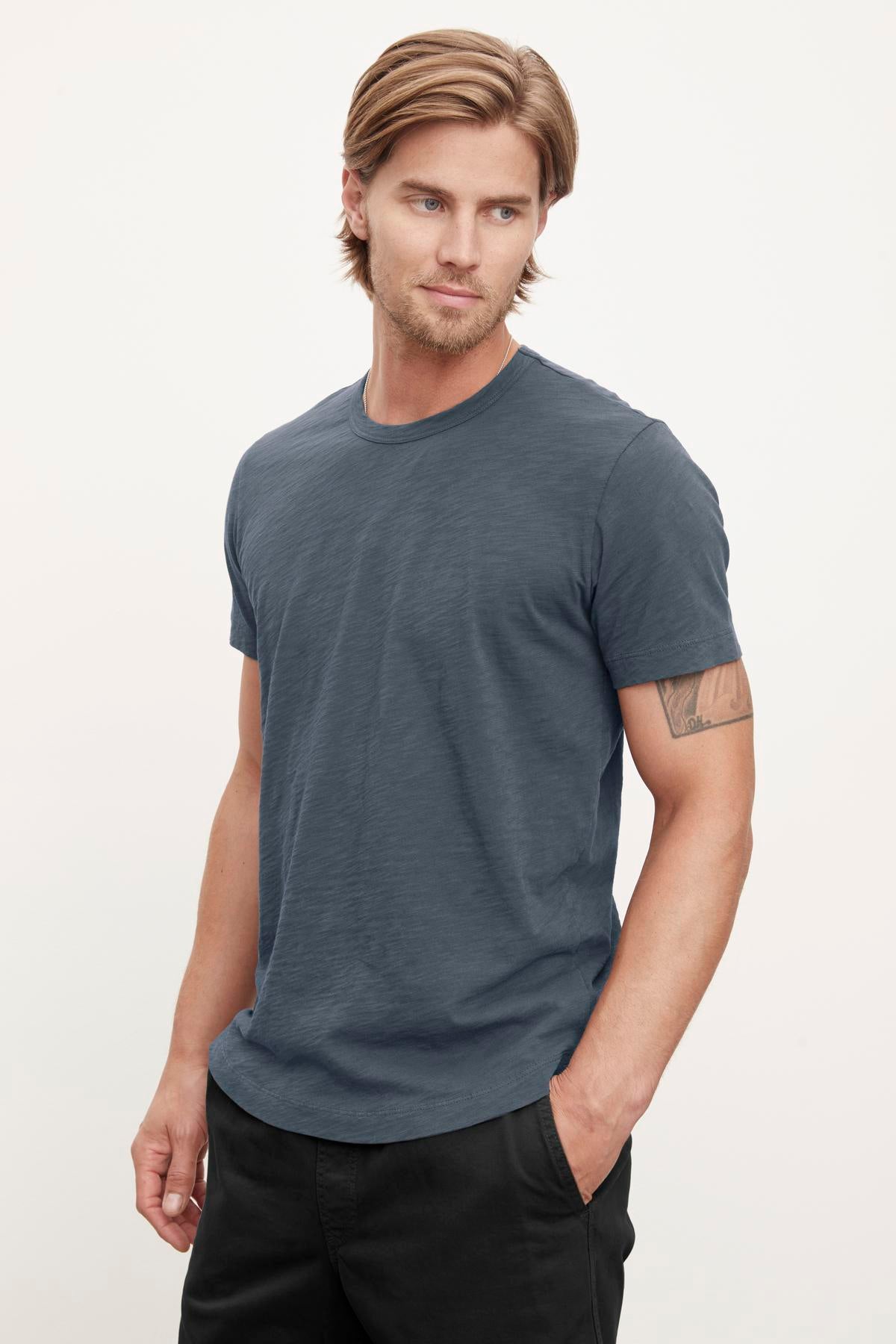   A man with medium-length hair wearing a blue Velvet by Graham & Spencer AMARO TEE and black pants stands with his left hand in his pocket, looking to his right, exuding a relaxed stylish feel. 