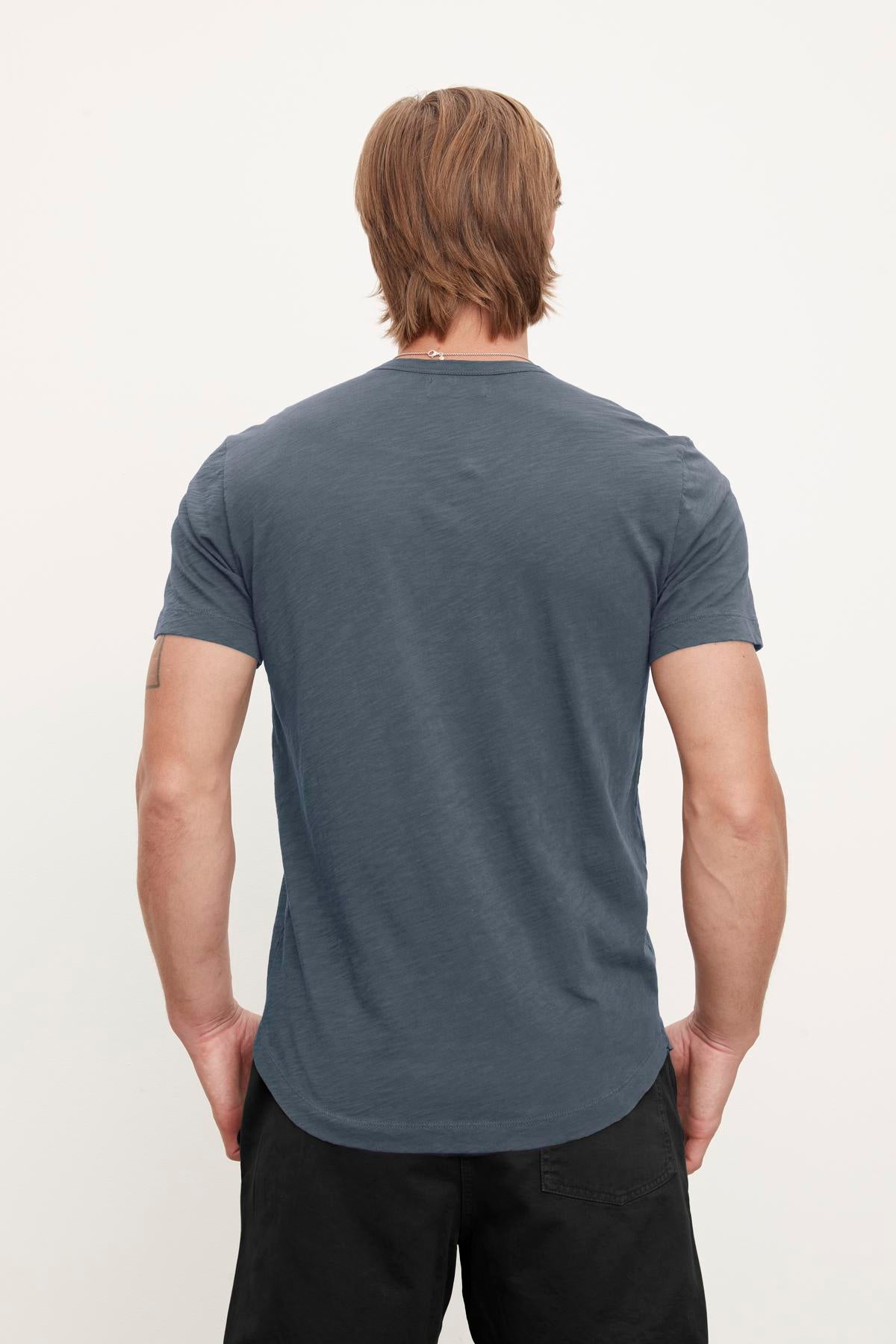   A person with shoulder-length hair is standing with their back to the camera, wearing a plain dark blue Velvet by Graham & Spencer AMARO TEE and black pants, exuding a relaxed stylish feel. 