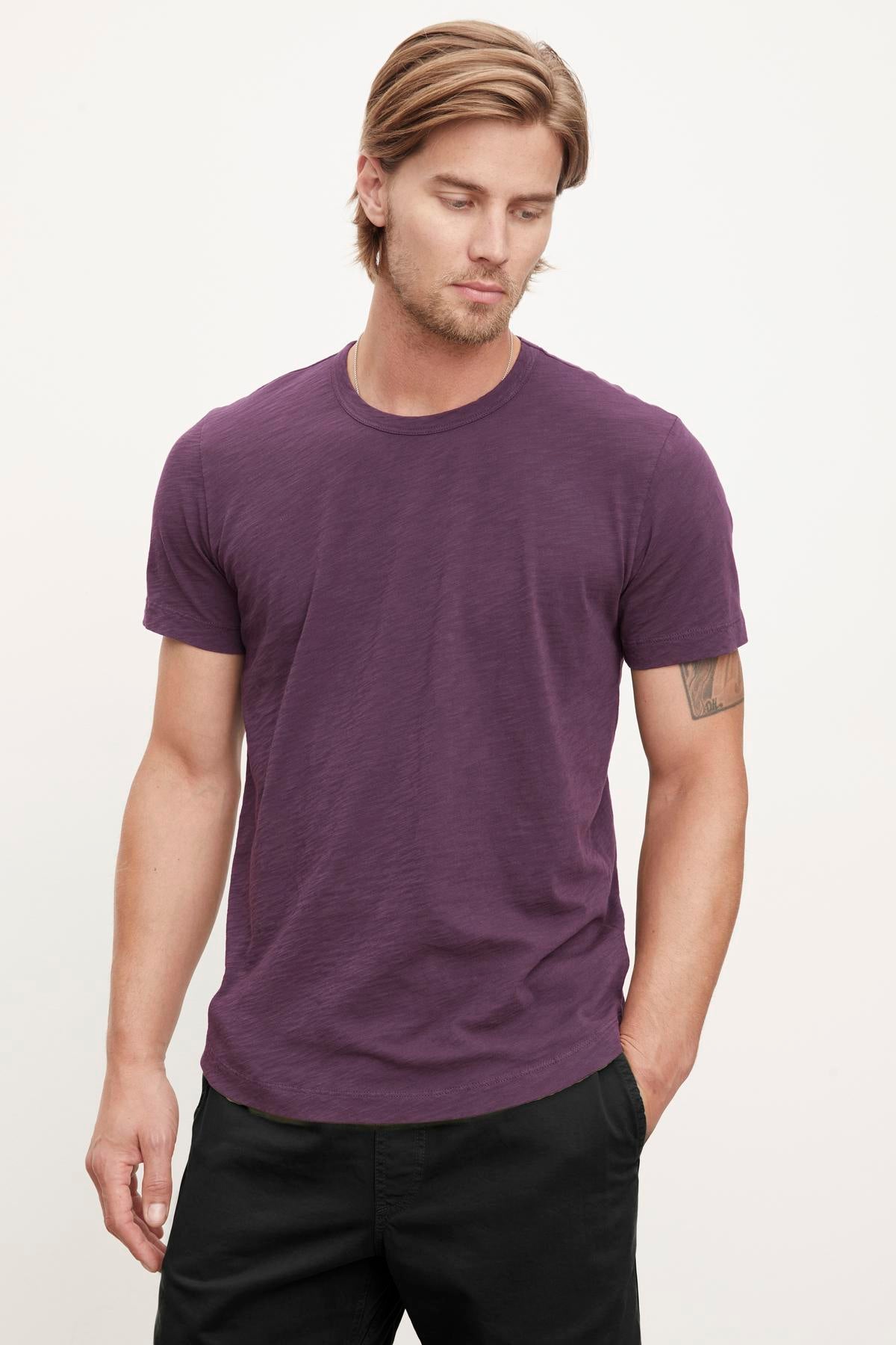   A person with light brown hair is wearing the AMARO TEE by Velvet by Graham & Spencer against a plain background, looking slightly downward. This purple t-shirt, featuring a classic crew neckline and crafted from lightweight Peruvian cotton, makes an excellent layering piece for any season. 