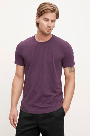 A person with light brown hair is wearing the AMARO TEE by Velvet by Graham & Spencer against a plain background, looking slightly downward. This purple t-shirt, featuring a classic crew neckline and crafted from lightweight Peruvian cotton, makes an excellent layering piece for any season.