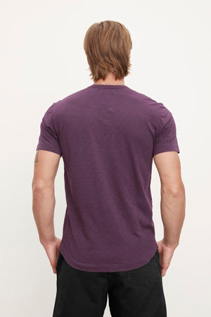 Man with medium-length hair wearing the Velvet by Graham & Spencer AMARO TEE, a lightweight Peruvian cotton purple T-shirt with a classic crew neckline, paired with black pants, facing away from the camera.