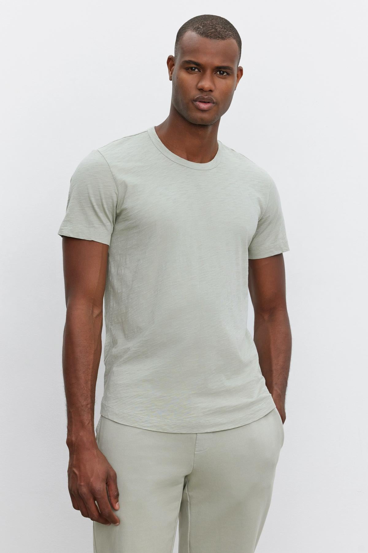   A person with a neutral expression stands against a plain white background, wearing a light grey AMARO TEE by Velvet by Graham & Spencer and matching grey pants, showcasing a versatile look with a relaxed and stylish feel. 