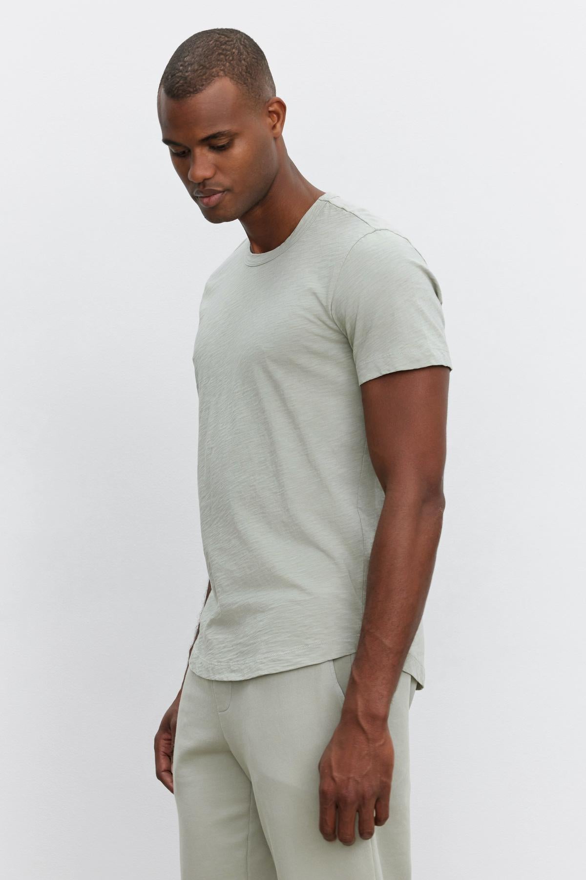   A man in a light grey Velvet by Graham & Spencer AMARO TEE and matching pants stands sideways against a plain white background, looking down, embodying a relaxed and stylish feel. 