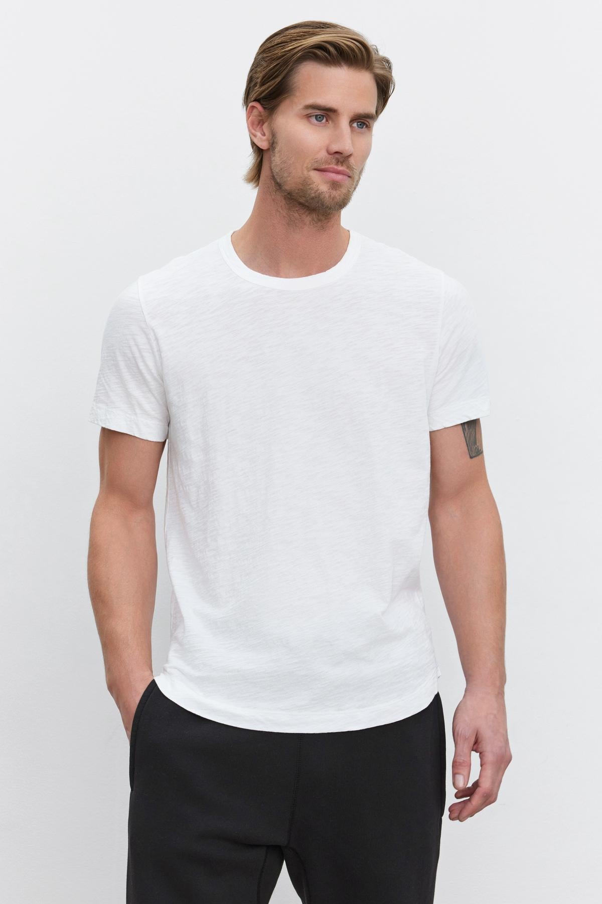   A man with blond hair posing in a white Velvet by Graham & Spencer slub knit AMARO TEE with a crew neck and black pants. 