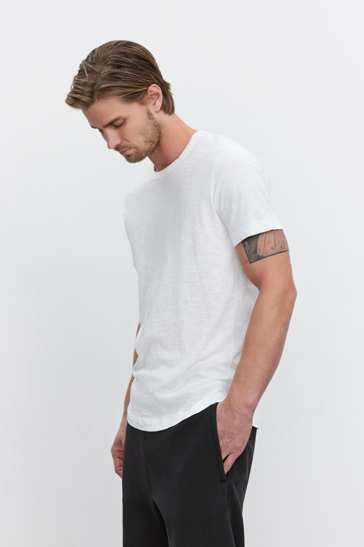   Man in a white crew neck Velvet by Graham & Spencer AMARO TEE with a heathered texture and black pants, looking downwards with a tattoo visible on his left arm. 