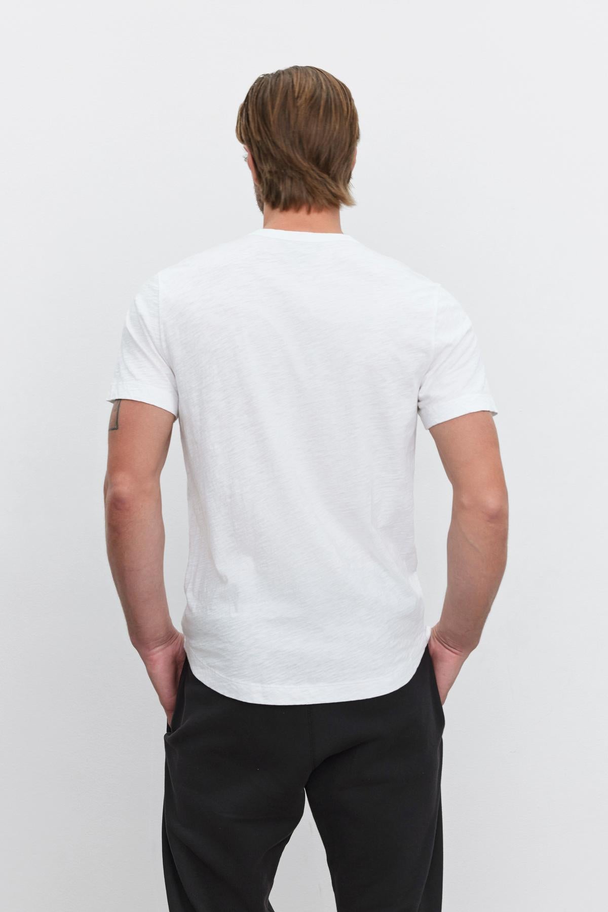   Man standing with his back to the camera wearing a white crew neck AMARO TEE with a heathered texture and black pants by Velvet by Graham & Spencer. 