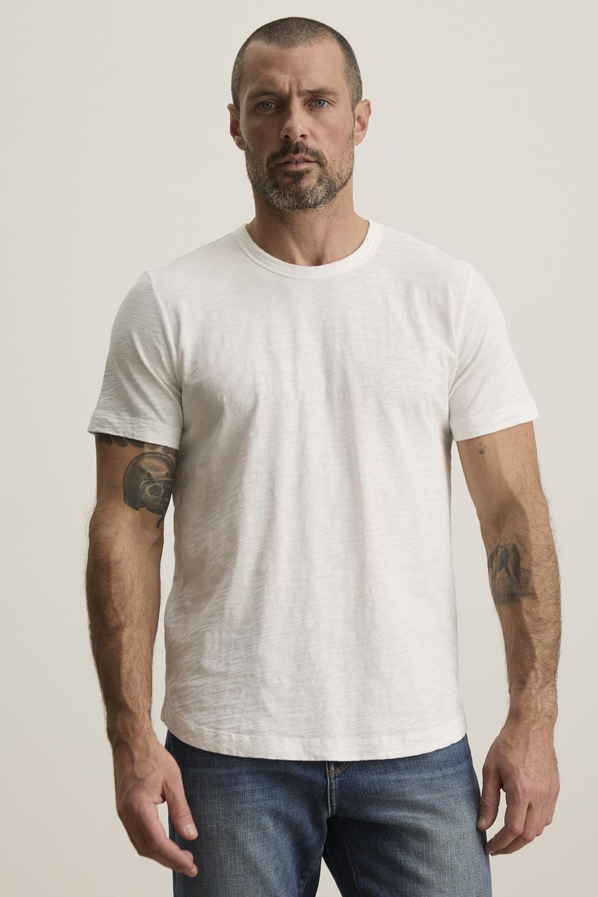   A person with short hair and a beard wears the versatile Velvet by Graham & Spencer AMARO TEE, made of Peruvian cotton, paired with jeans against a plain background. Its classic crew neckline makes it an ideal layering piece for any occasion. 