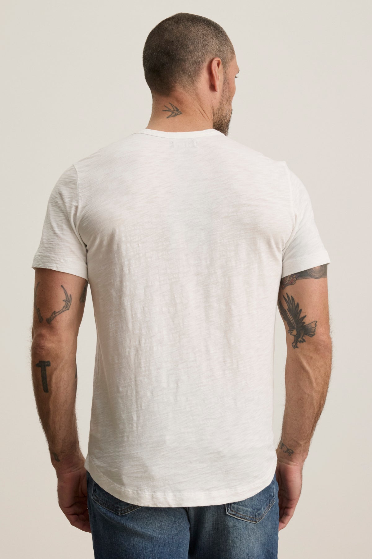   A man with short hair and several tattoos on his arms and neck is wearing a Velvet by Graham & Spencer AMARO TEE, a white crew-neckline shirt made of soft Peruvian cotton, along with blue jeans, facing away from the camera. 