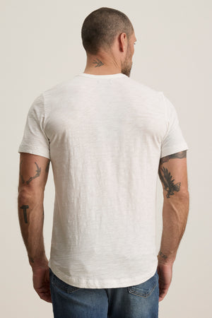 A man with short hair and several tattoos on his arms and neck is wearing a Velvet by Graham & Spencer AMARO TEE, a white crew-neckline shirt made of soft Peruvian cotton, along with blue jeans, facing away from the camera.