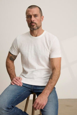 A man with a beard and short hair sits on a stool, wearing the AMARO TEE by Velvet by Graham & Spencer—a white Peruvian cotton T-shirt with a classic crew neckline—and blue jeans, looking at the camera. This versatile piece adds comfort and style to his relaxed look.