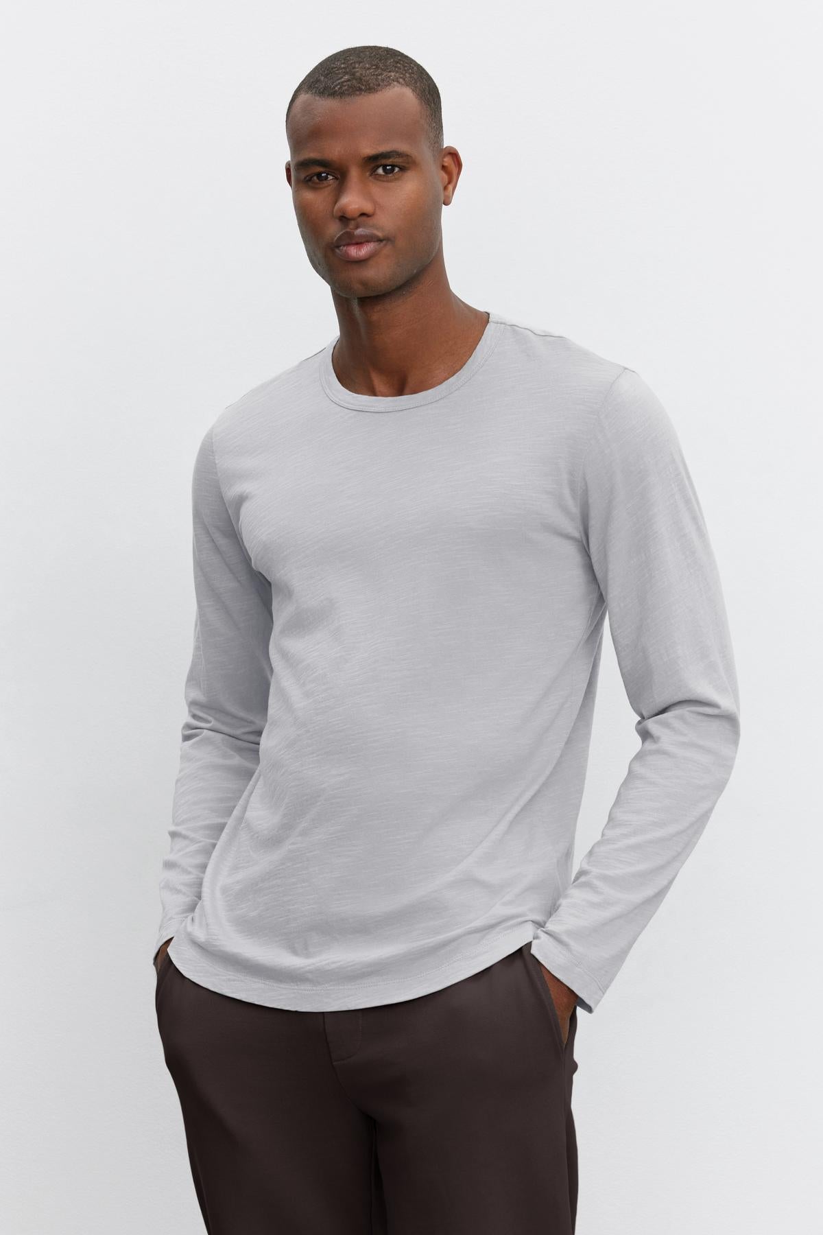 A man wearing the KAI TEE from Velvet by Graham & Spencer—a light gray long-sleeve shirt crafted from lightweight Peruvian cotton, featuring a classic crew neckline—and dark pants stands against a plain white background.-38264831803585