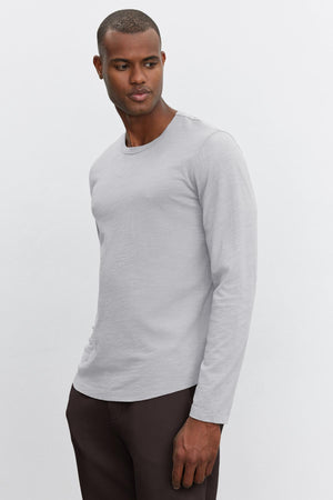 A person wearing the KAI TEE by Velvet by Graham & Spencer, a lightweight long-sleeve shirt made of Peruvian cotton in light gray, and dark pants stands against a plain white background, showcasing a timeless design.