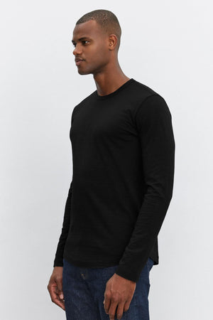 A man wearing a black KAI TEE long-sleeve shirt with a classic crew neckline by Velvet by Graham & Spencer and blue jeans stands against a plain white background.