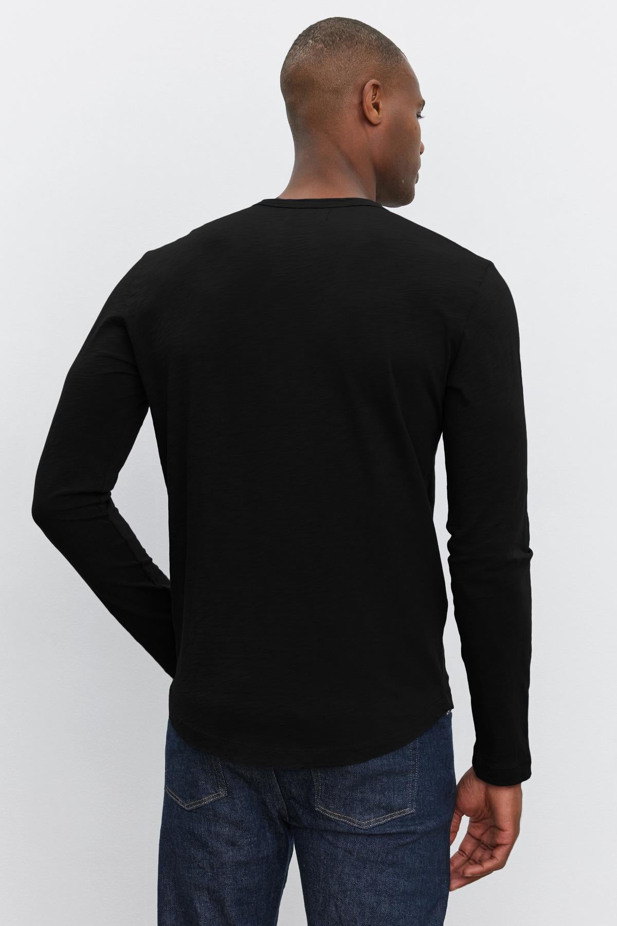 A person standing with their back to the camera, wearing a black long-sleeve KAI TEE from Velvet by Graham & Spencer featuring a classic crew neckline and dark jeans.-37469162275009