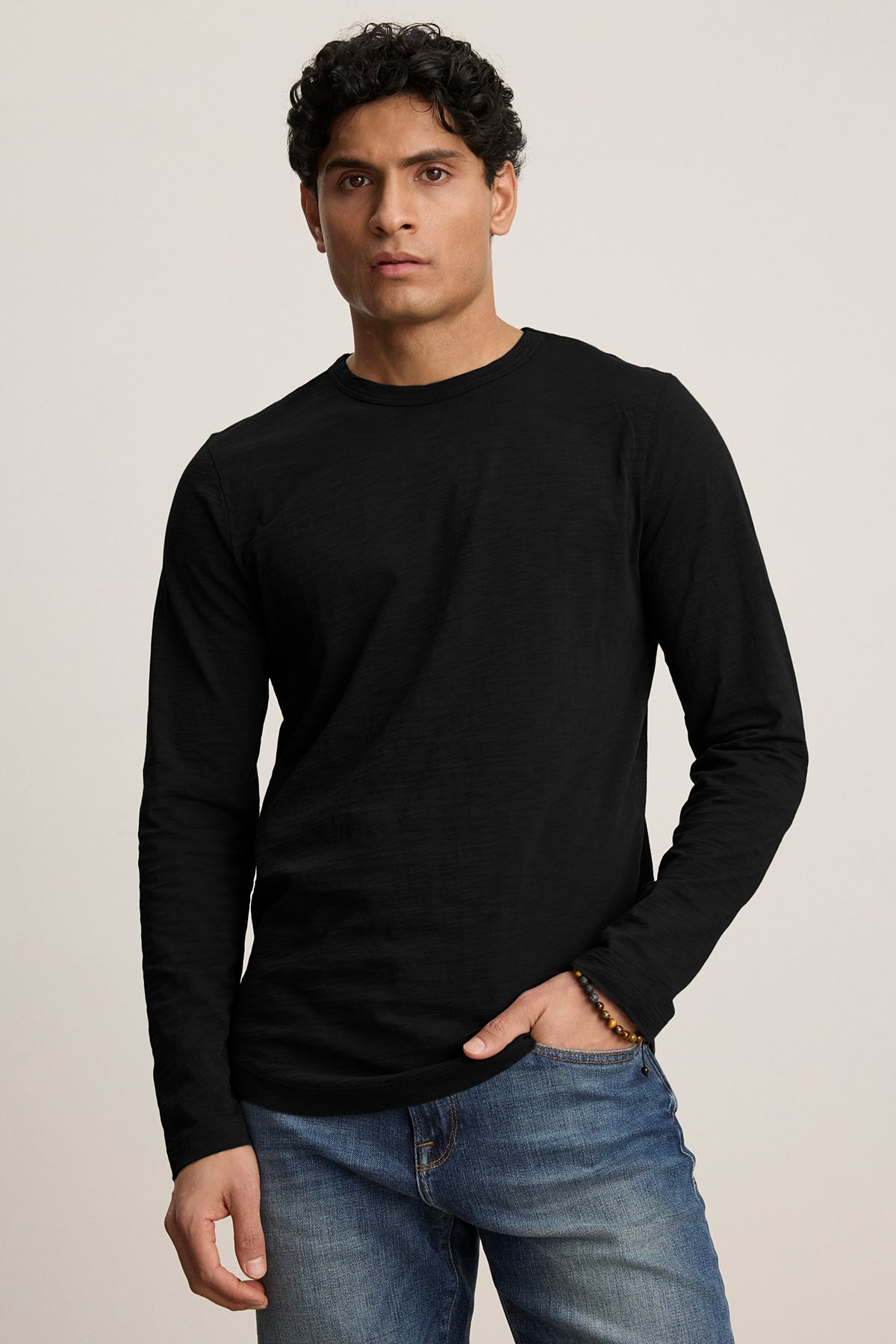   A curly-haired individual in a KAI TEE, a black long-sleeve shirt with a crew neckline by Velvet by Graham & Spencer, wears blue jeans and stands casually with one hand in their pocket. 