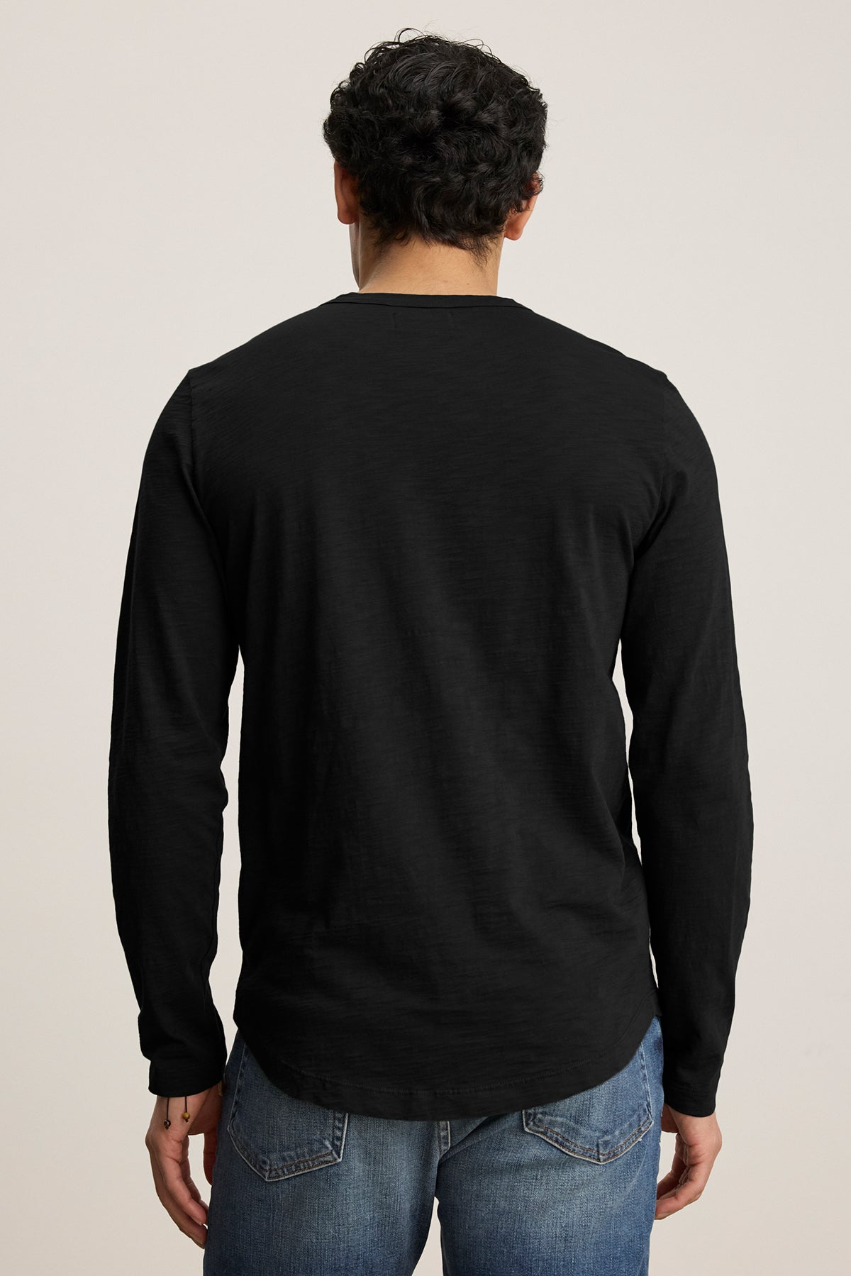   Back view of a person with curly hair wearing the KAI TEE by Velvet by Graham & Spencer, a black long-sleeve shirt with a classic crew neckline, paired with jeans and set against a plain background. 