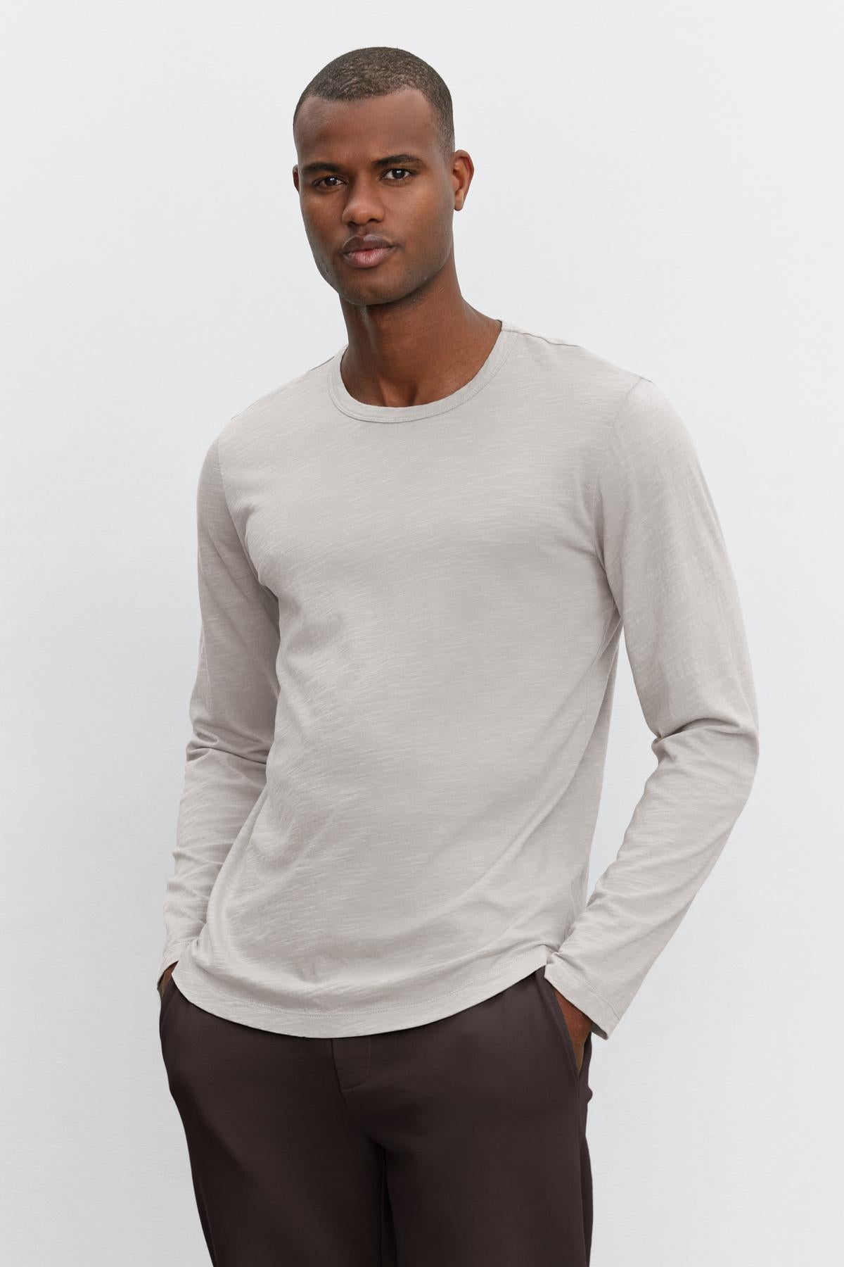   A person wearing the KAI TEE by Velvet by Graham & Spencer, a light gray long-sleeve shirt crafted from lightweight Peruvian cotton with a classic crew neckline, stands in front of a plain white background while paired with dark pants. The shirt features a timeless design. 