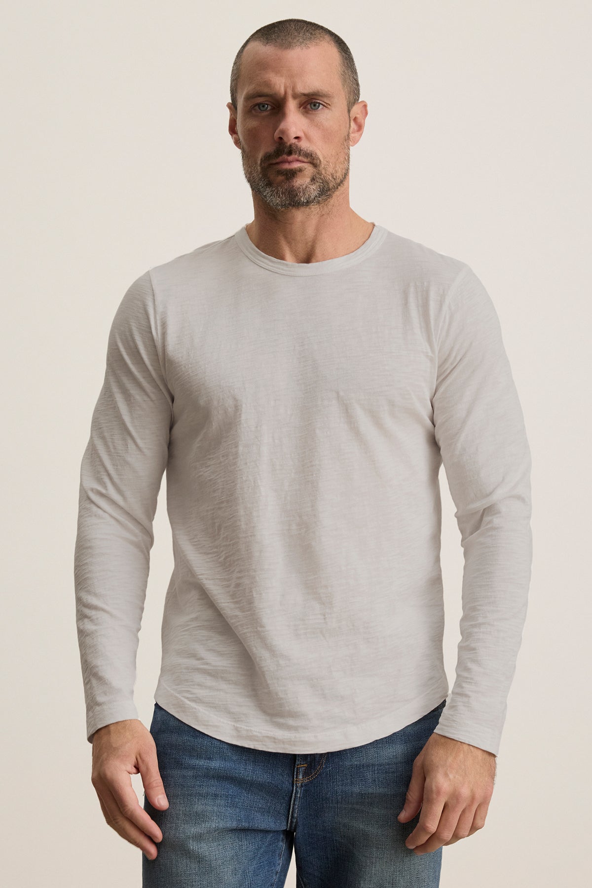   A bearded man in a KAI TEE by Velvet by Graham & Spencer, a light gray long-sleeve Peruvian cotton shirt, and blue jeans stands against a plain background. 