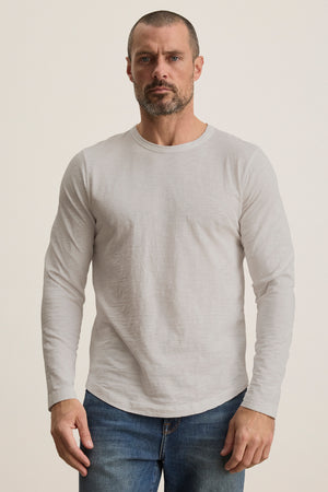 A bearded man in a KAI TEE by Velvet by Graham & Spencer, a light gray long-sleeve Peruvian cotton shirt, and blue jeans stands against a plain background.