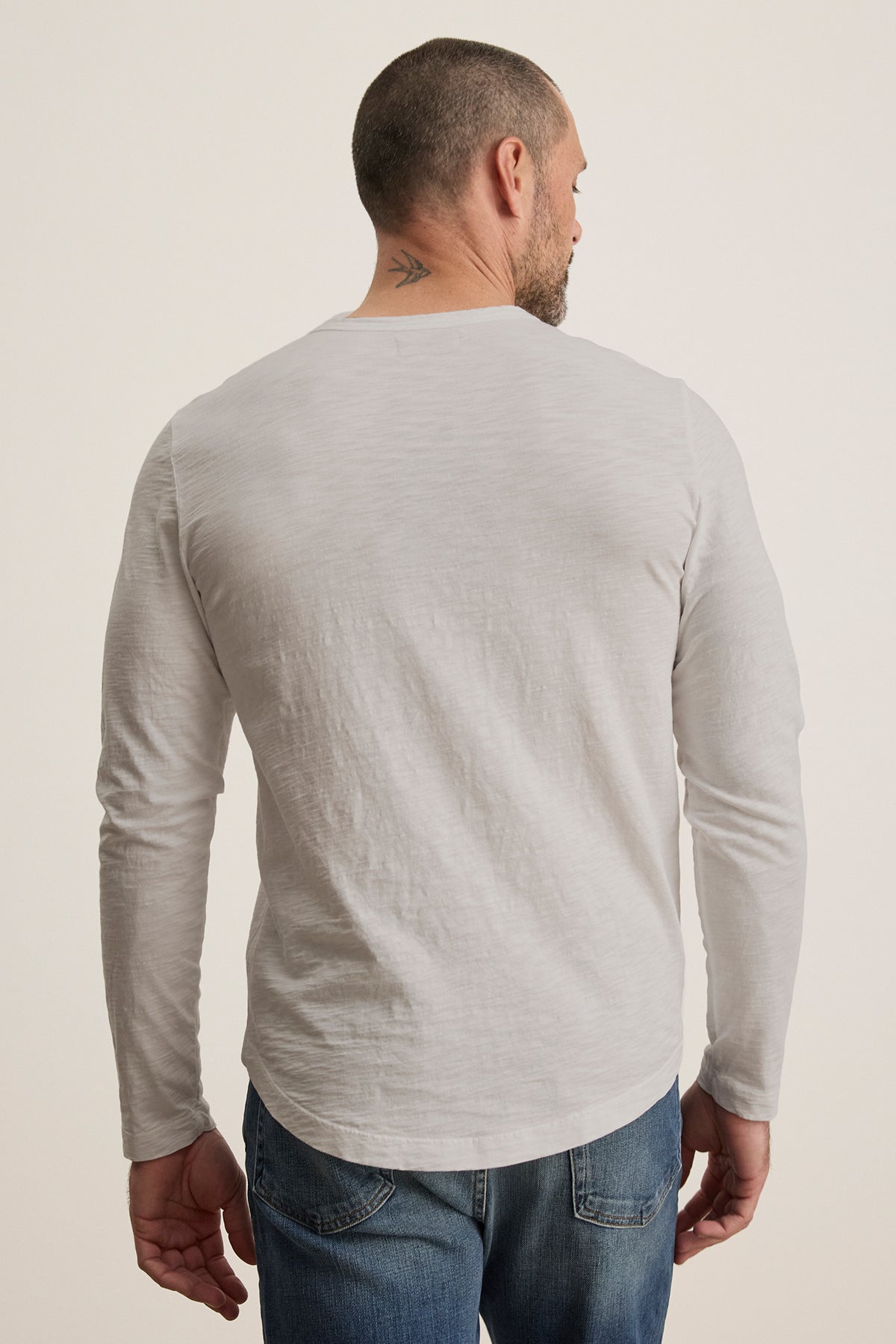 A man with short hair and a small neck tattoo, dressed in a KAI TEE by Velvet by Graham & Spencer—a white long-sleeve shirt with a crew neckline and timeless design—paired with jeans, is facing away from the camera.-38807449469121