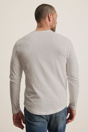 A man with short hair and a small neck tattoo, dressed in a KAI TEE by Velvet by Graham & Spencer—a white long-sleeve shirt with a crew neckline and timeless design—paired with jeans, is facing away from the camera.