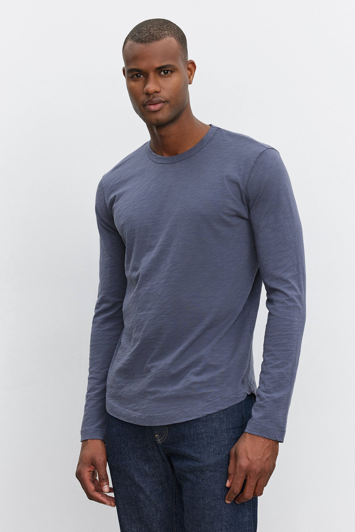 A man stands against a plain background, wearing a long-sleeve, dark grey KAI TEE from Velvet by Graham & Spencer and jeans.-37893903220929