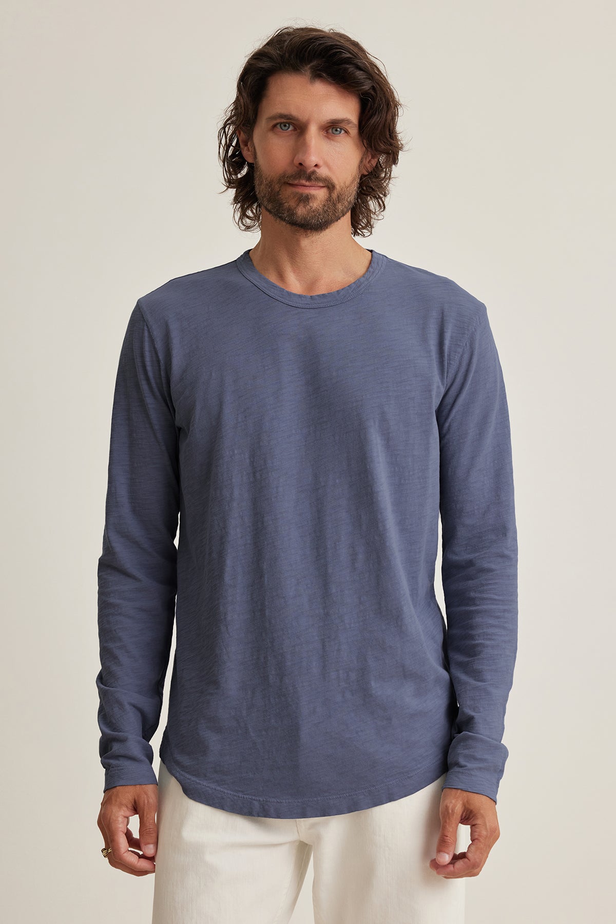 Wearing a KAI TEE by Velvet by Graham & Spencer, made of soft Peruvian cotton, a person with shoulder-length hair stands against a neutral background. The blue long-sleeve shirt, paired with white pants, features a classic crew neckline for a timeless look.-38807449075905