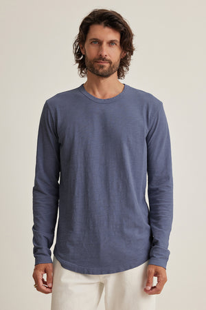 Wearing a KAI TEE by Velvet by Graham & Spencer, made of soft Peruvian cotton, a person with shoulder-length hair stands against a neutral background. The blue long-sleeve shirt, paired with white pants, features a classic crew neckline for a timeless look.