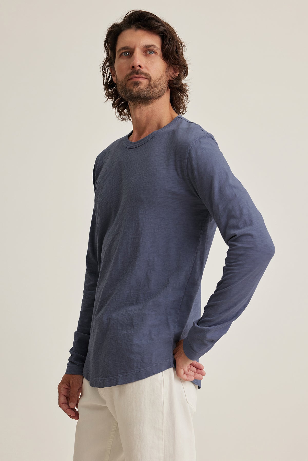   A person with long hair in the KAI TEE by Velvet by Graham & Spencer, crafted from Peruvian cotton, and white pants stands against a plain background, gazing slightly upwards. 