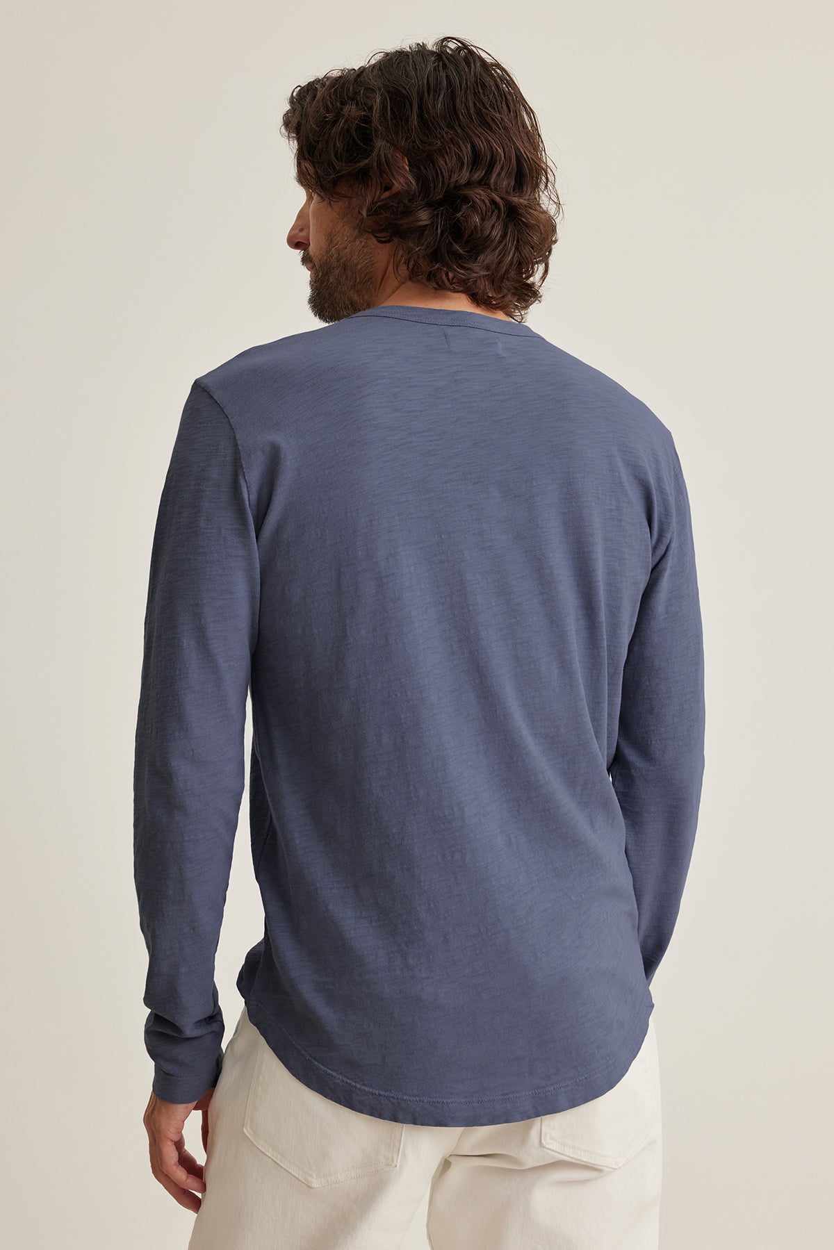   A person with shoulder-length hair wears the Velvet by Graham & Spencer KAI TEE, a timeless blue long-sleeve shirt made from Peruvian cotton, paired with white pants. They're facing away from the camera against a neutral background. 