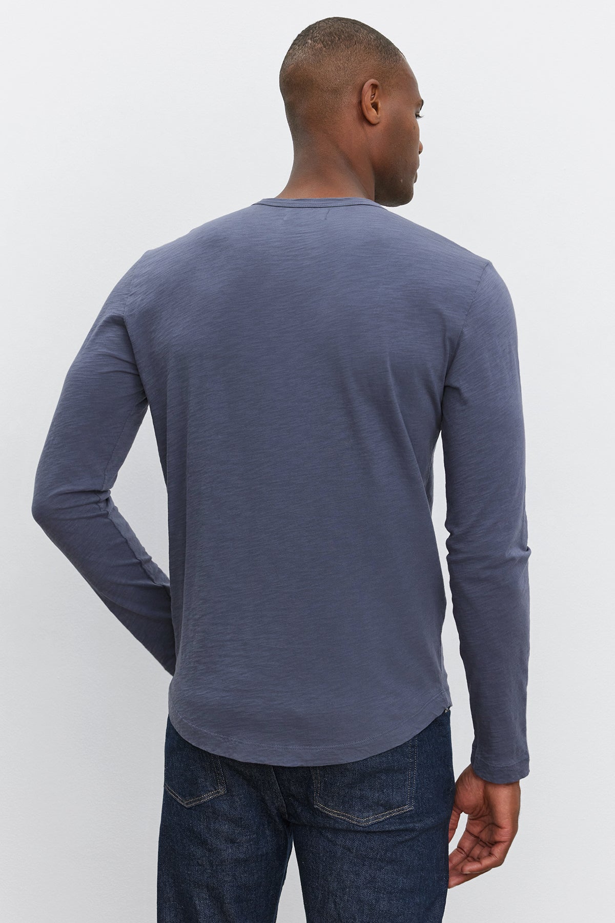   A man is standing with his back to the camera, wearing the Velvet by Graham & Spencer KAI TEE, a long-sleeved dark gray shirt with a classic crew neckline, paired with dark blue jeans against a light-colored background. 