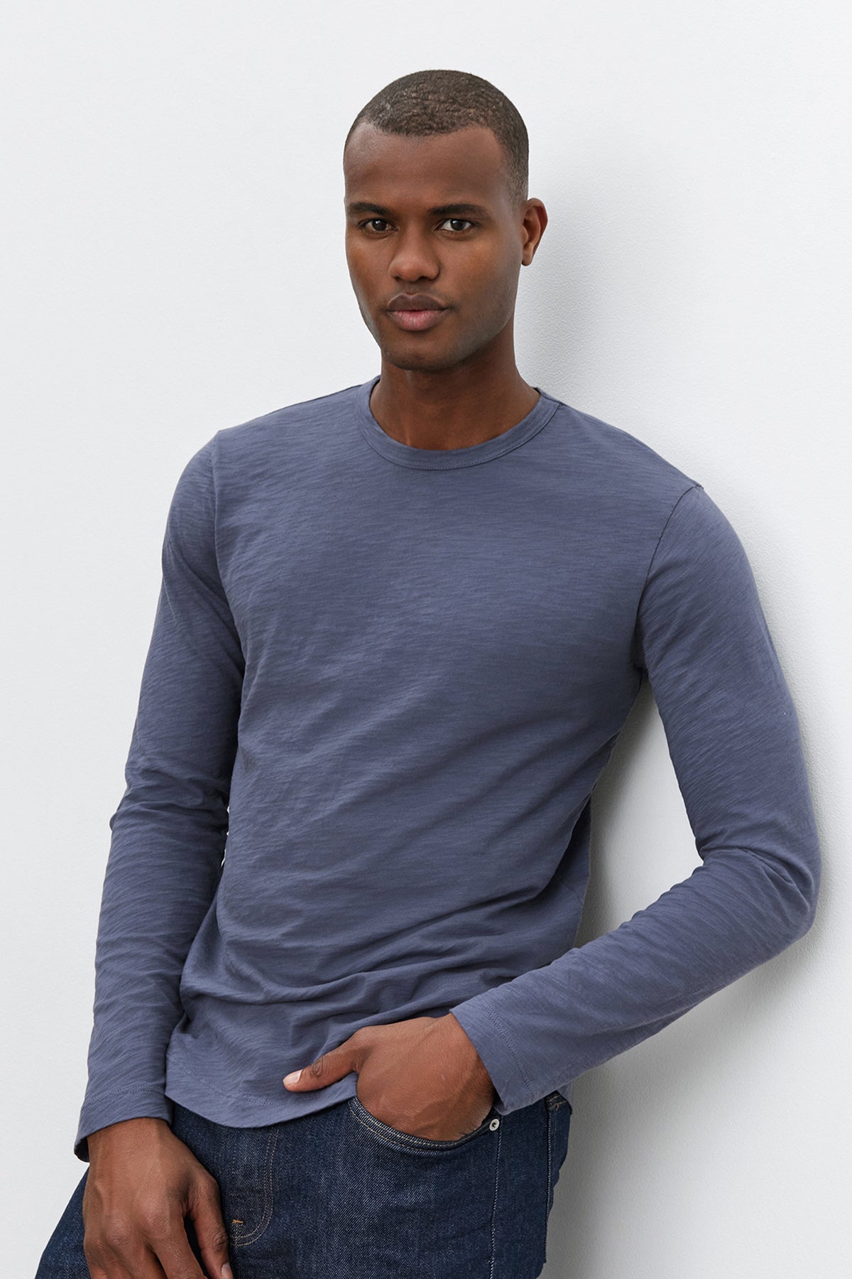   A man wearing Velvet by Graham & Spencer's KAI TEE, a long-sleeved blue shirt with a classic crew neckline, and dark jeans stands against a plain, white background. With one hand in his pocket, he effortlessly showcases the timeless design of his attire. 