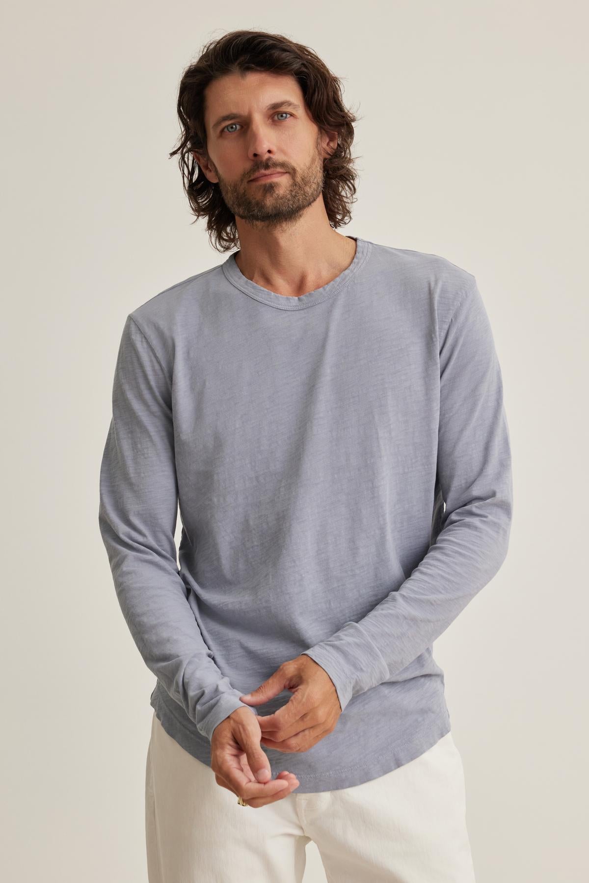 A man with shoulder-length hair and a beard wears the KAI TEE by Velvet by Graham & Spencer—a light gray long-sleeve shirt with a classic crew neckline—and white pants, standing against a plain background.-38643308101825