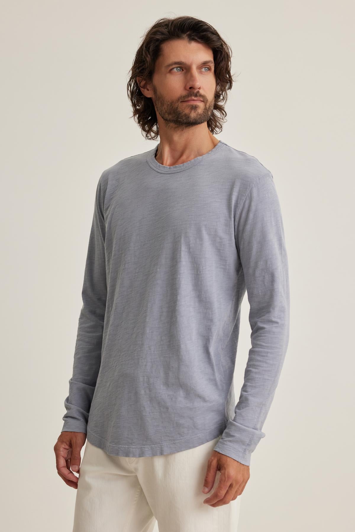   Wearing Velvet by Graham & Spencer's KAI TEE, featuring a gray long-sleeve and classic crew neckline, paired with white pants, a person stands against a plain background, gazing to the side. 