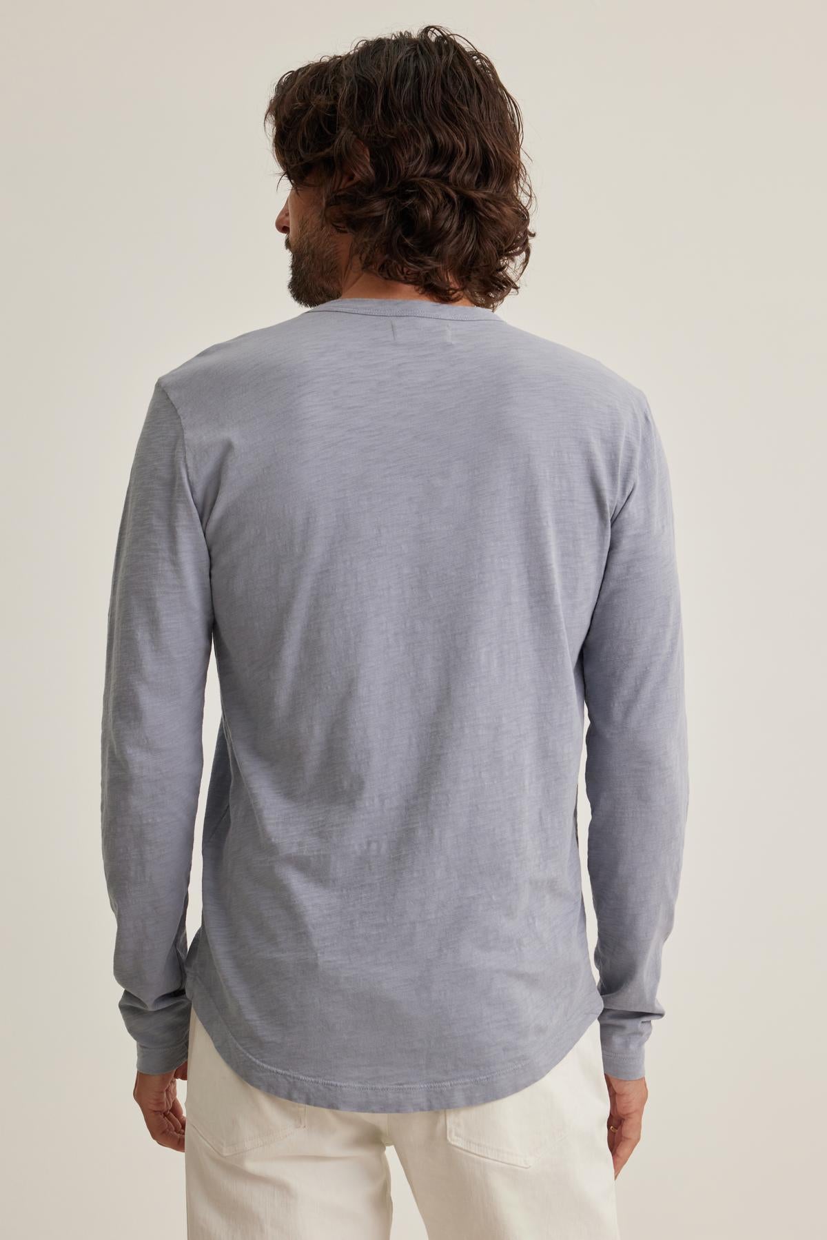   A person with long hair, seen from behind, dons the KAI TEE by Velvet by Graham & Spencer—a classic crew neck shirt in light gray—paired with white pants against a plain background, embodying timeless design. 