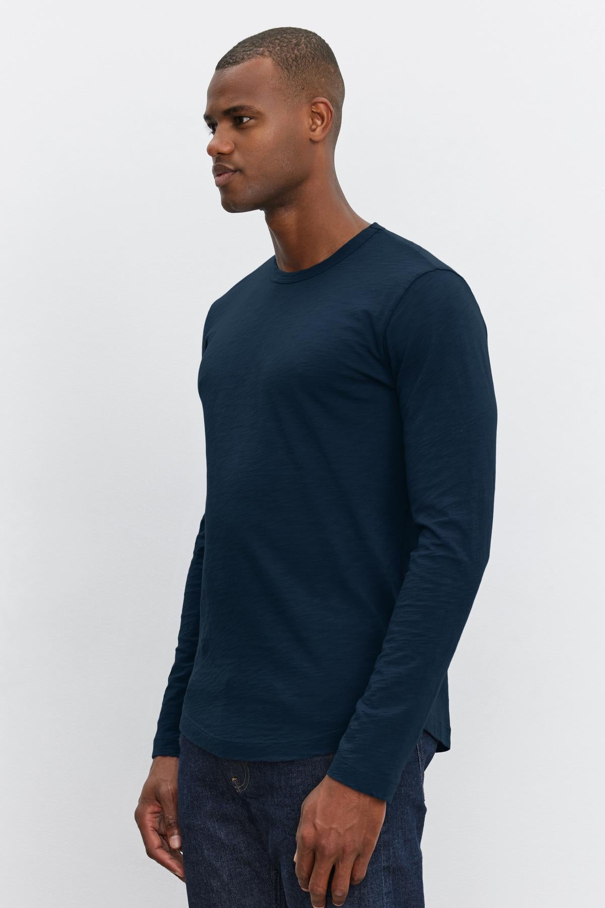   A man wearing a plain, long-sleeve, dark blue KAI TEE by Velvet by Graham & Spencer with a modern curved hemline and dark pants stands against a plain white background. 