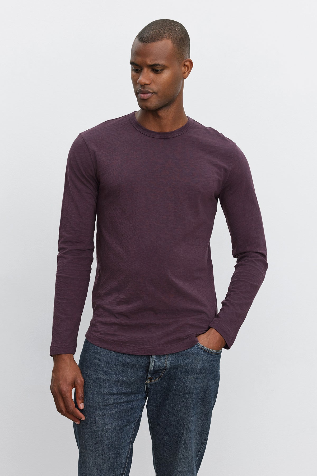   A person stands against a plain background, wearing the KAI TEE by Velvet by Graham & Spencer – a long-sleeved, plum-colored shirt made of Peruvian cotton – paired with blue jeans. With one hand in their pocket, they are looking downwards. 