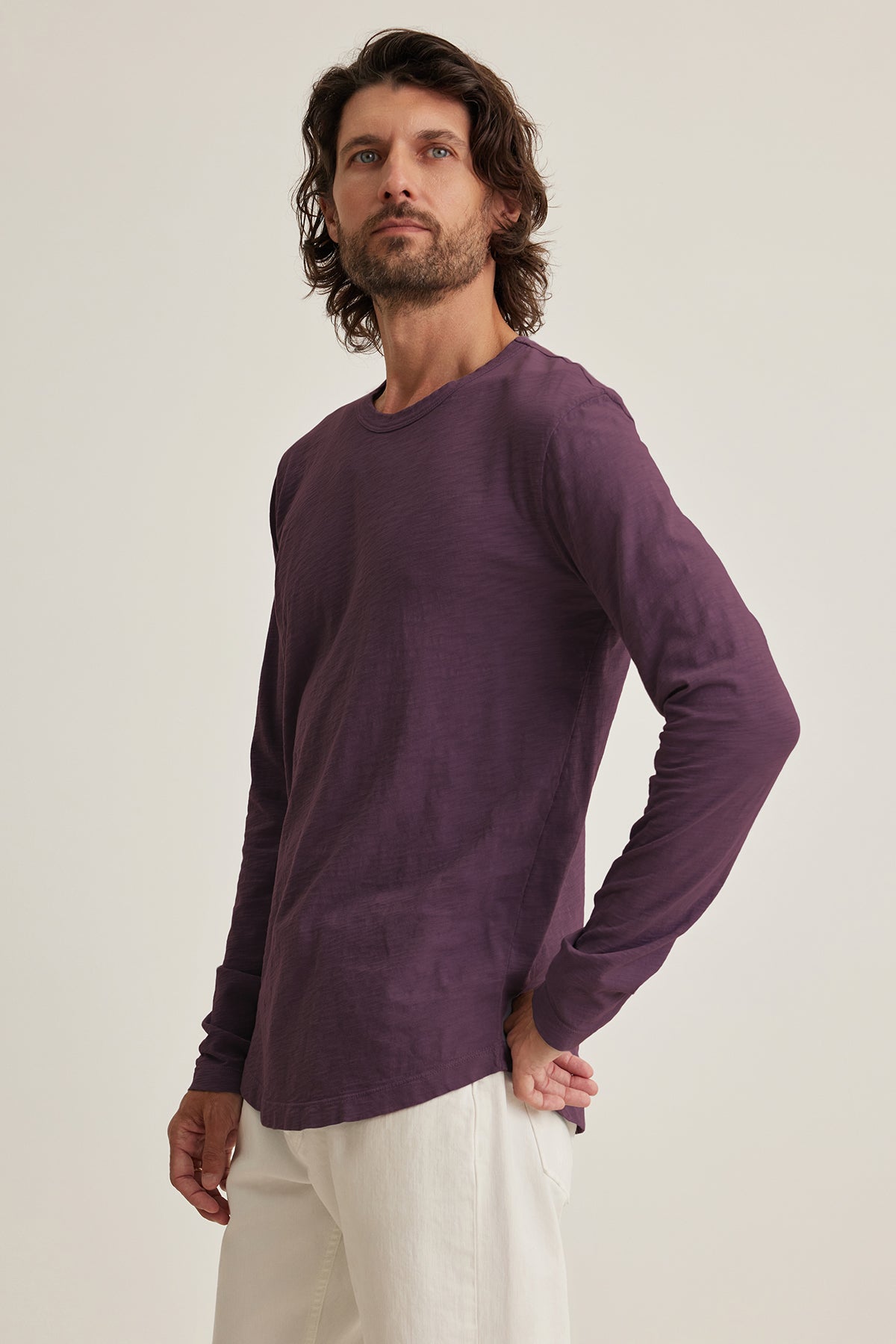   A man with long hair wears Velvet by Graham & Spencer's KAI TEE, a purple long-sleeve shirt paired with white pants, exuding timeless design as he gazes slightly upward against a plain background. 