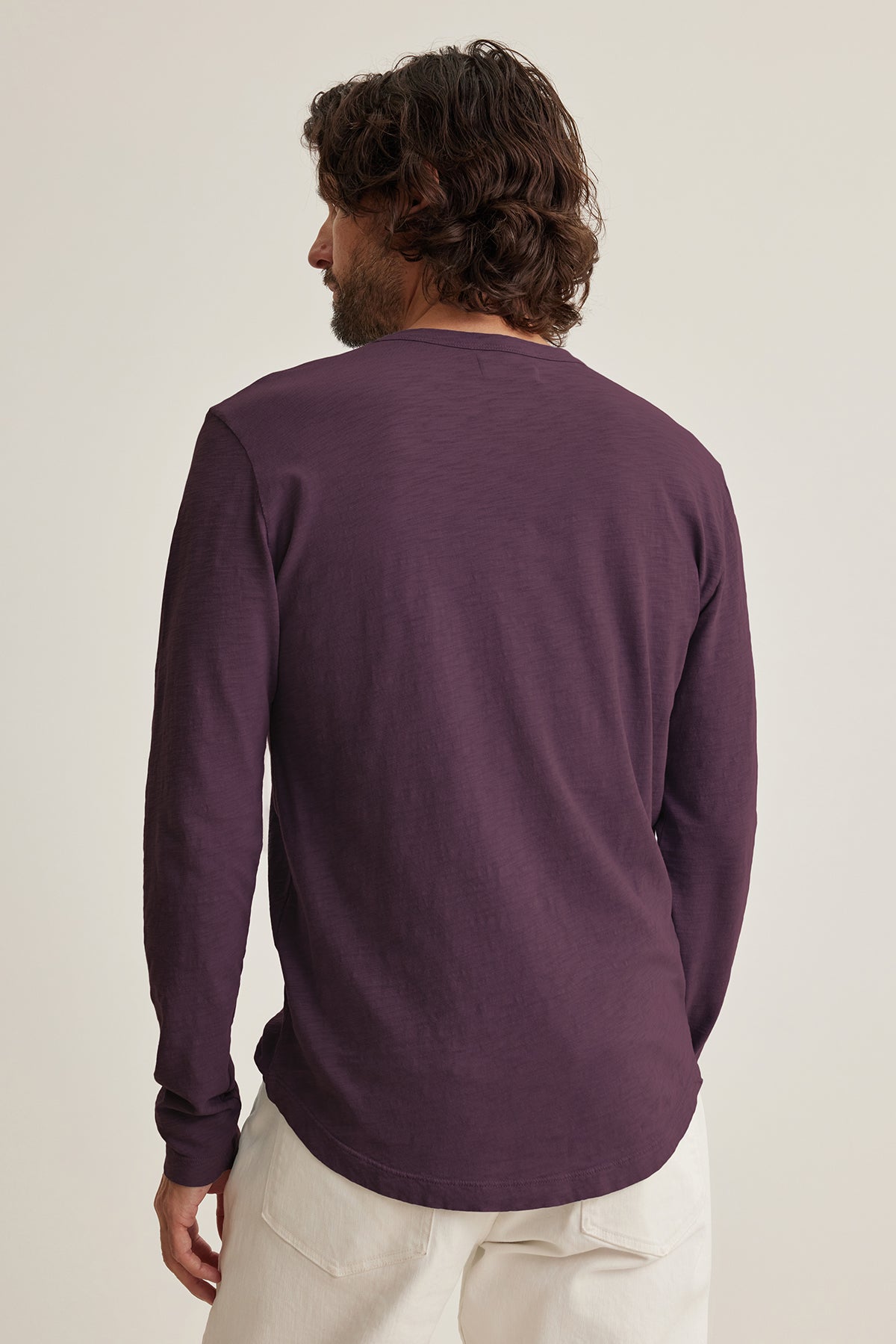   A dark-haired man wears a maroon KAI TEE by Velvet featuring a crew neck, paired with light pants, and is facing away. 