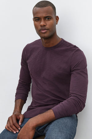 A person wearing the long-sleeve KAI TEE shirt by Velvet by Graham & Spencer, made from Peruvian cotton, and blue jeans sits against a plain white background.