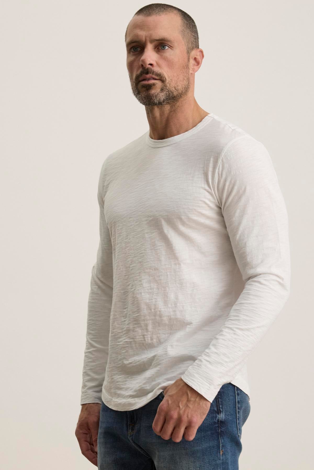   A man with a beard stands against a plain background, wearing jeans and the KAI TEE by Velvet by Graham & Spencer, a white long-sleeve shirt featuring a classic crew neckline made of luxurious Peruvian cotton. 